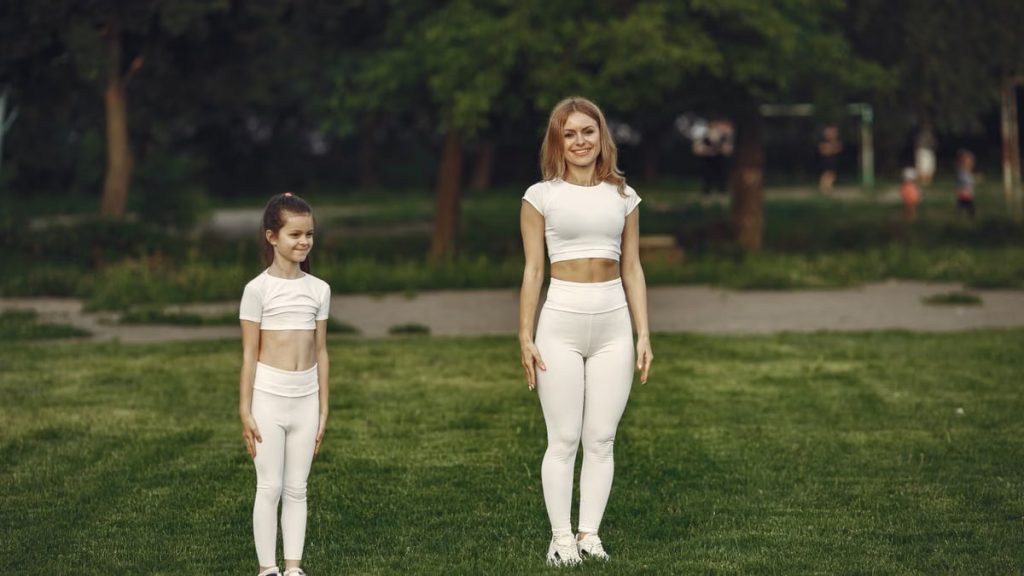 A woman and girl in white activewear standing on a grass field outdoors, smiling.
