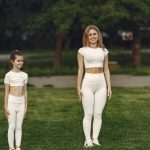 A woman and girl in white activewear standing on a grass field outdoors, smiling.