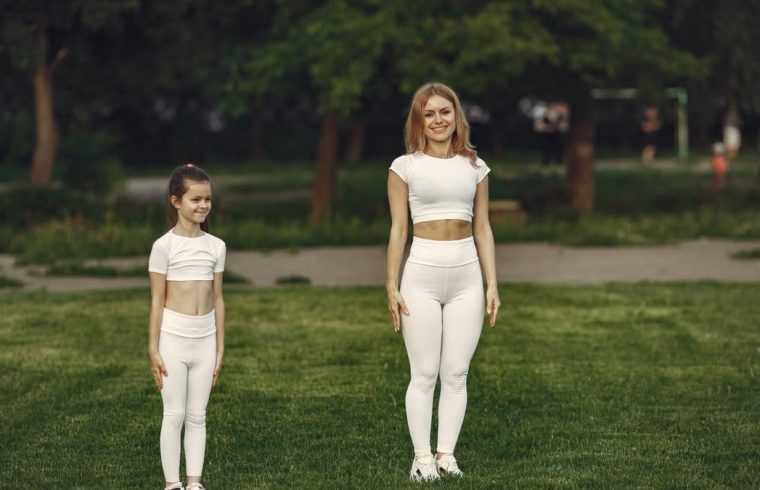 A woman and girl in white activewear standing on a grass field outdoors, smiling.